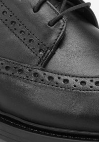 Scarpa in pelle nera con dettagli brogue, caratterizzata da perforazioni lungo le cuciture, chiusura con lacci e superficie liscia.