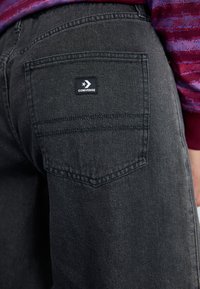 Jeans de mezclilla negros con un bolsillo trasero que presenta una etiqueta rectangular del logo de Converse. Detalles de textura y costura sutiles visibles.