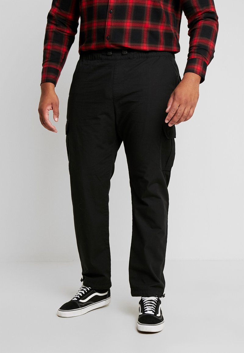 Schwarze Cargo-Hose mit elastischem Bund und Seitentaschen, getragen mit schwarz-weißen Sneakers. Über sichtbar ist ein kariertes Hemd in Rot und Schwarz.