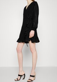 Svart klänning med v-ringning, texturerad tyg, långa puffärmar, volangkant och figurnära midja. Bärs med svarta klacksandaler.