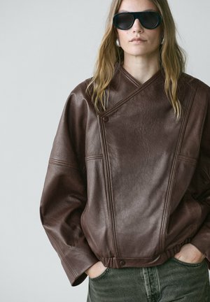 Femme portant une grande veste en cuir marron, lunettes de soleil foncées, boucles d'oreilles en perles, mains dans les poches, sur un fond gris clair uni.