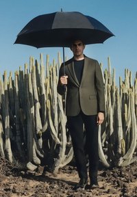 Mężczyzna w dopasowanej oliwkowozielonej marynarce i czarnych spodniach trzyma czarny parasol. Otoczony wysokimi, zielonymi kaktusami na tle jasnego nieba.