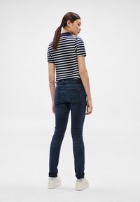 Prúžkované tričko s krátkym rukávom v kombinácii s tmavomodrými džínsami, ktoré majú priliehavý strih, a svetlými teniskami, pohľad zozadu.