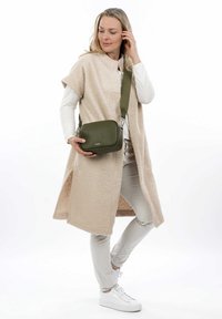 Gilet in pile beige a maniche corte sopra una maglia bianca a maniche lunghe, abbinato a pantaloni chiari e sneakers bianche. Tieni in mano una piccola borsa a tracolla verde.