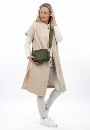 Gilet in pile beige a maniche corte sopra una maglia bianca a maniche lunghe, abbinato a pantaloni chiari e sneakers bianche. Tieni in mano una piccola borsa a tracolla verde.