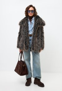 Palton din blană artificială cu dungi gri și maro, purtat peste o cămașă din denim și un pulover gri, asortat cu blugi deschiși și cizme scurte maro închise, ținând o geantă maro.