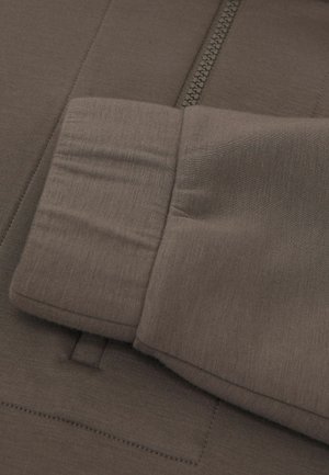 Braunes, zum Schließen mit Reißverschluss versehenes Hemd mit glatter Textur, dezentem Glanz und Manschette mit genähten Details. Der Reißverschluss verläuft vertikal in der Mitte.