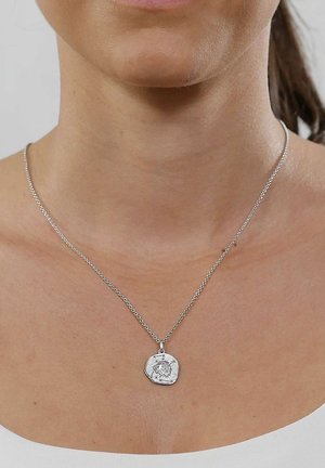 Close-up of a woman's neck wearing a silver chain necklace with a round pendant featuring an engraved lion head design.