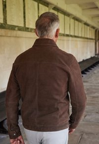 Chaqueta de ante marrón con un cuello estructurado, costuras detalladas y un diseño ajustado, vista desde la espalda frente a un fondo de pared de piedra.