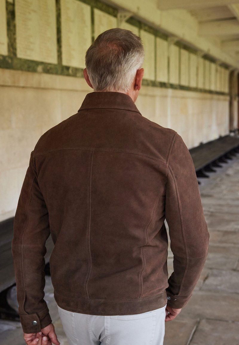 Chaqueta de ante marrón con un cuello estructurado, costuras detalladas y un diseño ajustado, vista desde la espalda frente a un fondo de pared de piedra.