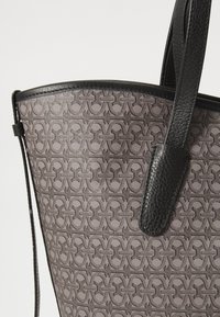 Grå mønstret tote bag med sorte læderkanter og -stropper. Har en glat overflade og et gentagende logo-design på stoffet.