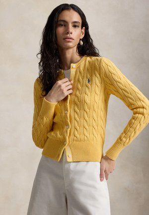 Hosszú sötét hajú nő sárga, kötött kardigánban és fehér nadrágban, semleges bézs háttér előtt áll.