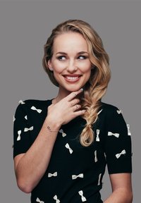 Vrouwelijk model gekleed in een zwart shirt versierd met kleine witte strik applicaties, met lang golvend blond haar in een losse vlecht gestyled. Zilveren armband gedragen.