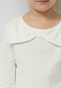 Kind trägt ein langärmliges weißes Oberteil mit einer großen Stoffschleife am Ausschnitt, die von den Schultern bis zu den Lippen sichtbar ist.