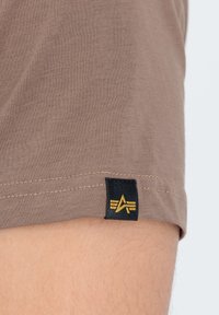 Camiseta corta marrón con una etiqueta negra que presenta un logo amarillo. La tela es suave y lisa, con costuras visibles en el dobladillo.