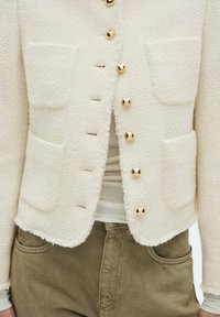 Veste texturée crème avec boutons dorés, deux poches avant et bords effilochés, associée à un haut clair et un pantalon vert olive.