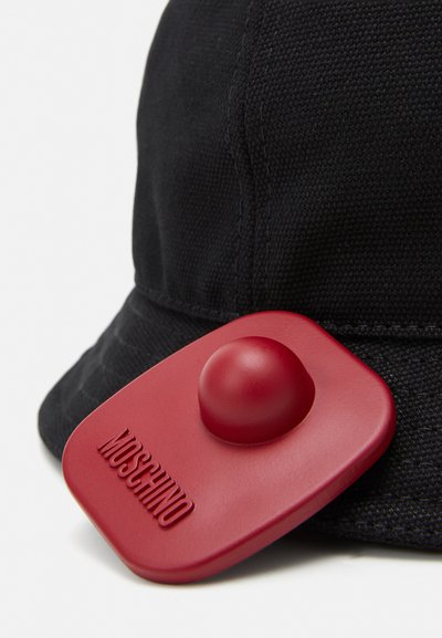 Pălărie bucket din material negru, cu un accent circular roșu neted pe care este embossat textul "MOSCHINO", evidențiind o suprafață texturată și o formă structurată.