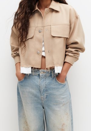 Personne aux cheveux longs et bouclés portant une veste courte beige sur une chemise blanche et un jean bleu ample, les mains dans les poches.