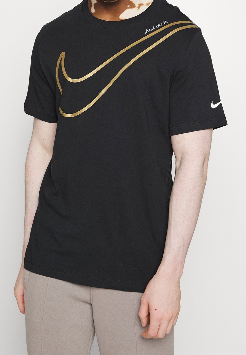 Osoba v černém tričku Nike s velkým zlatým swoosh a nápisem "Just do it." na hrudi, v kombinaci s béžovými kalhotami.