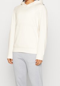 Persona che indossa una felpa con cappuccio color crema con tasca frontale e pantaloni grigio chiaro, in piedi di fronte a uno sfondo neutro.