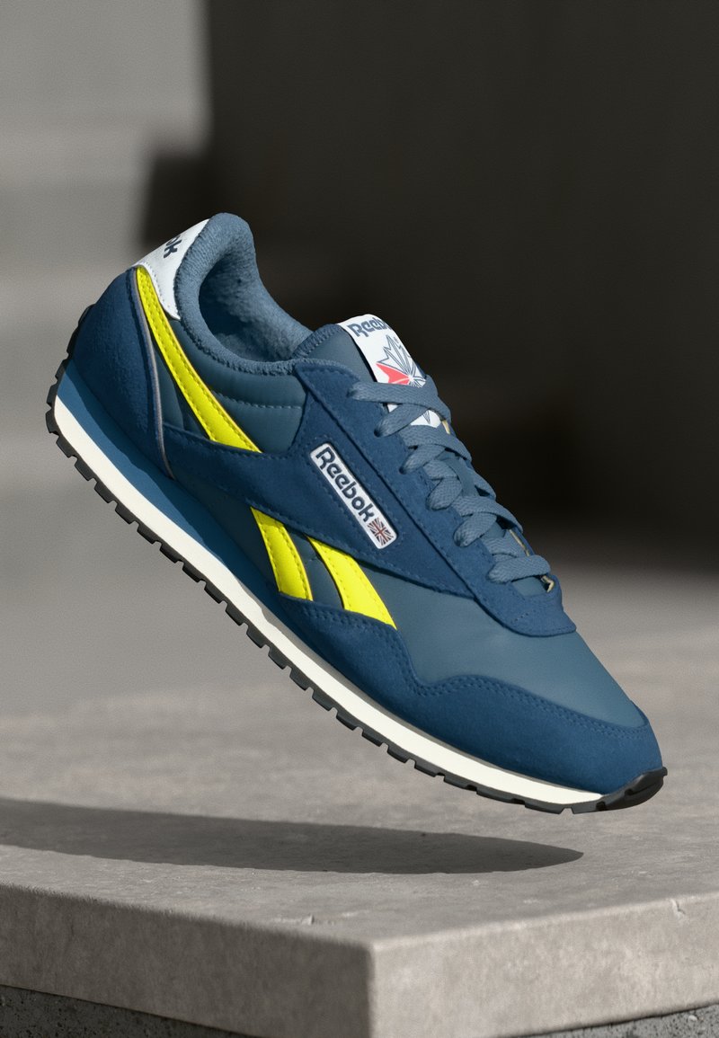 Reebok sportbačiai su tamsiai mėlynu viršumi ir geltonais akcentais, lygių odos ir zomšos medžiagų derinys bei tekstūruotas juodas guminis padas.