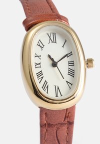 Gulltonet oval klokke med hvit skive, svarte romerske tall og brun teksturert lærreim. Har en krone på høyre side.