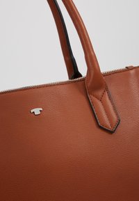 Sac à main en cuir marron avec deux poignées, fermeture éclair argentée et petit logo argenté en forme de T sur surface texturée.