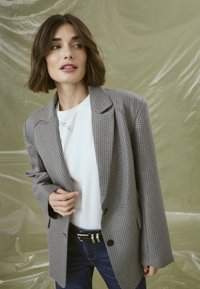 Šedý strukturovaný blazer s velkými klopami, na dvě knoflíky, kombinovaný s bílou košili a tmavými džínami. Hladká látka s jemným vzorem.