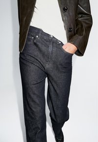 Jeans in denim blu scuro con vestibilità rilassata, caratterizzati da uno stile a cinque tasche, bottoni in metallo e cuciture a contrasto, abbinati a una giacca di pelle nera.