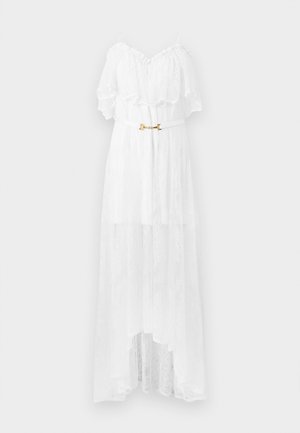 Femme portant une robe blanche en dentelle à taille haute et basse, avec bretelles spaghetti et sandales noires à talons hauts, debout devant un fond uni.