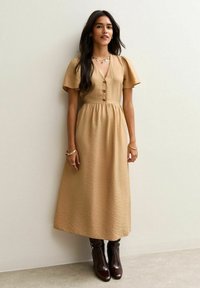Robe midi beige avec des manches courtes bouffantes, un devant à boutons et une taille froncée. Portée avec des bottes marron montantes. Texture lisse, motif subtil.