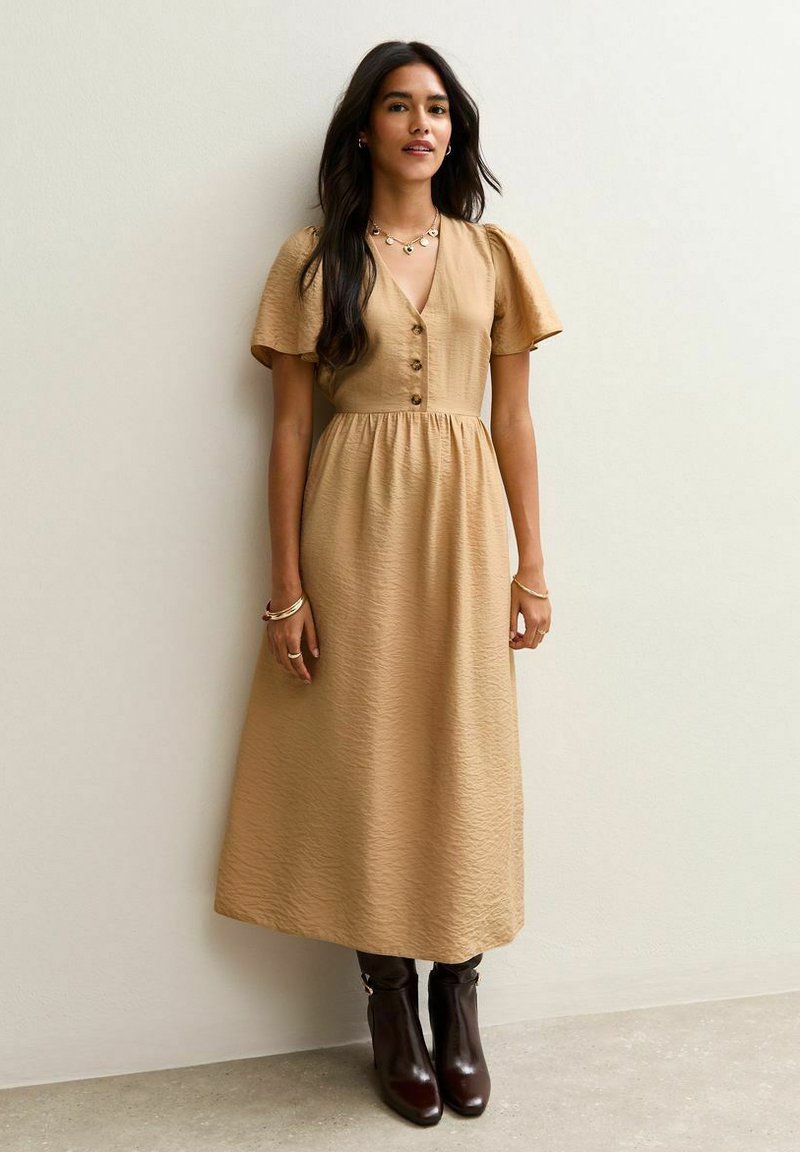 Robe midi beige avec des manches courtes bouffantes, un devant à boutons et une taille froncée. Portée avec des bottes marron montantes. Texture lisse, motif subtil.