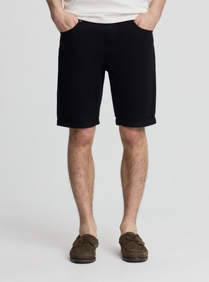 Mężczyzna w czarnych szortach z podwiniętymi nogawkami, białej koszuli i brązowych mokasynach, stojący z rękami w kieszeniach na jasnoszarym tle.