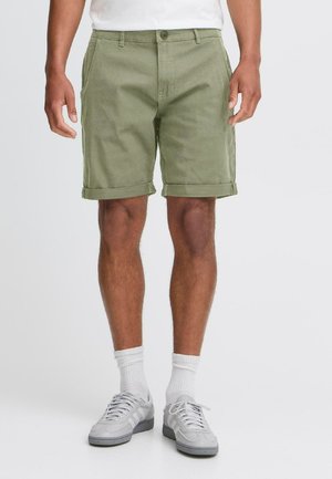 Pantalones cortos de algodón verde oliva con dobladillos enrollados, bolsillos frontales y cierre de un solo botón, combinados con zapatillas grises y calcetines blancos.