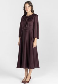 Bordowa satynowa sukienka z okr ągłym dekoltem, długimi rękawami i przewężoną talią, sięgająca do długości midi. Połączona z czerwonymi szpilkami.