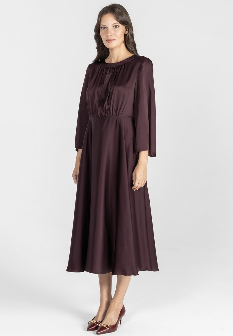 Bordowa satynowa sukienka z okrągłym dekoltem, długimi rękawami i przewężoną talią, sięgająca do długości midi. Połączona z czerwonymi szpilkami.