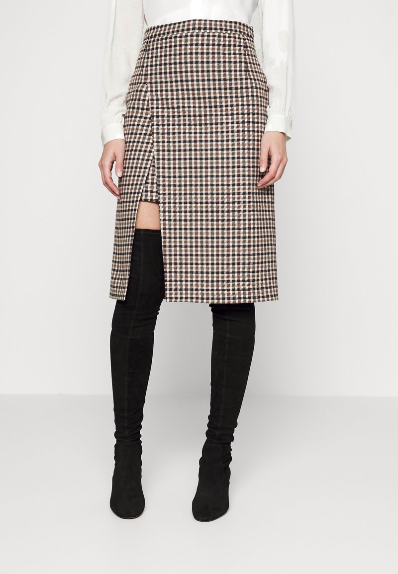 Donna che indossa una gonna plaid fino al ginocchio con spacco davanti, abbinata a stivali neri alti fino alla coscia e a una maglietta bianca a maniche lunghe.