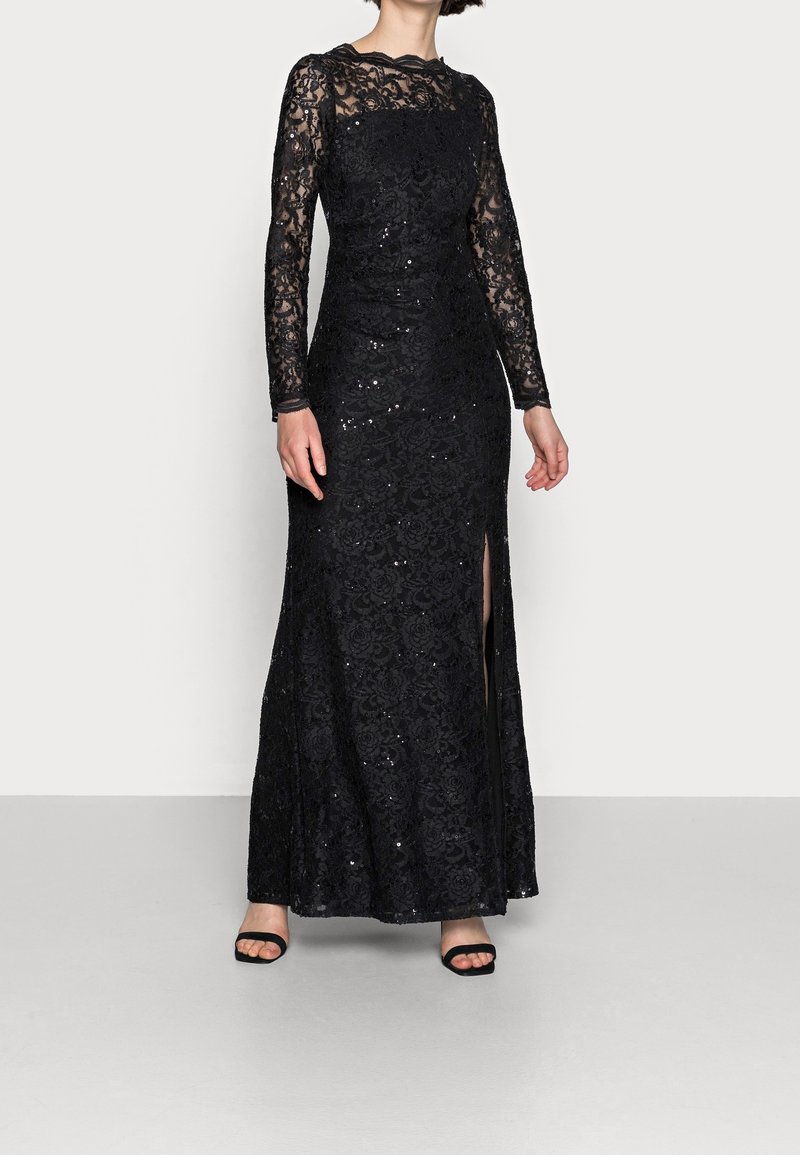 Abito lungo in pizzo nero con paillettes, silhouette aderente, motivo floreale e spacco alla coscia. Indossato con tacchi neri.