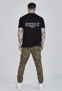 Čierne tričko s krátkym rukávom s výrazným bielym logom značky na chrbte, doplnené zelenými maskáčovými cargo nohavicami a bielymi teniskami.