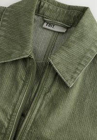 Zipzáras dzseki texturált olívazöld anyagból, galléros nyakkivágással. Finom csíkos mintázattal és belső béléssel rendelkezik.