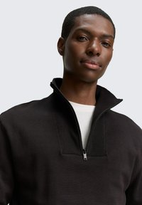 Zwarte pullover met een rits tot halverwege de kraag, ribbels in de stof en een gladde textuur. Heeft een witte binnenlaag eronder.