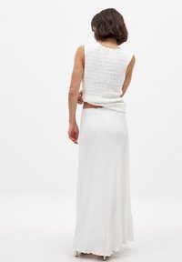 Lühikeste pruunide juustega naine, kandes valget tekstuuriga ilma varrukateta toppi ja pikka seelikut, seisab seljaga kaamera poole tavalises valges ruumis.