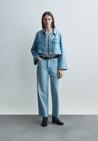 Tânără stând în picioare, purtând o jachetă scurtă din denim deschis la culoare, blugi cu talie înaltă și curea neagră, top gri și ghete negre până la gleznă pe podea gri.