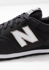 Svart sportskor med mesh tyg och syntetiska detaljer. Har en framträdande vit "N"-logotyp. Hållbar gummisula och texturerade snören.