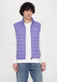 Colete puffer roxo claro com colarinho em pé, fecho frontal e logótipo EA7. Usado sobre uma camisa de manga longa branca, combinado com jeans azuis.