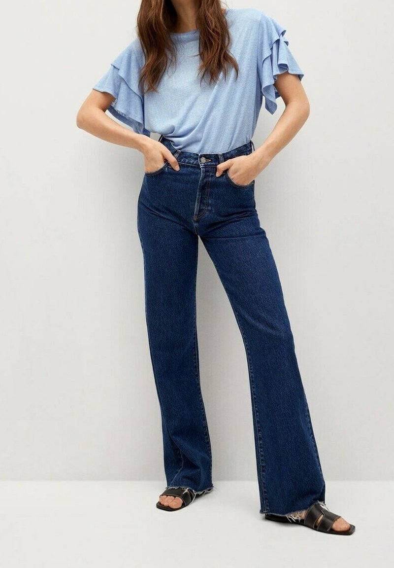 Lysblå kortærmet top med rynkede ærmer, parret med højtaljede mørkeblå denim flared jeans. Sandalerne har brede stropper og flad sål.