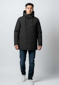 Jeune homme portant un manteau d'hiver noir à capuche, un jean bleu et des baskets blanches, se tenant devant un fond clair uni.