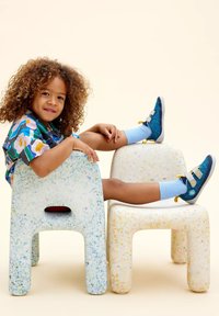 Dwa kolorowe, nakrapiane plastikowe taborety w bieli i kremie. Dziecko siedzi, mając na sobie niebieskie skarpetki i buty z żółtymi akcentami, uśmiechając się.