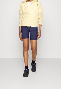 Ljusgul zip-jacka med huva, marinblå shorts och svarta skor. Jackan har dragsko och textilåta; shortsen har en enkel design.