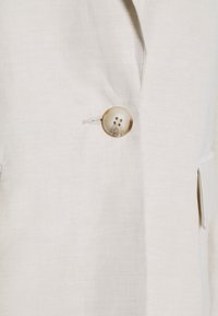 Veste en lin beige clair avec un grand bouton rond à quatre trous, poches latérales et coupe ajustée. Texture lisse avec peu de plis.